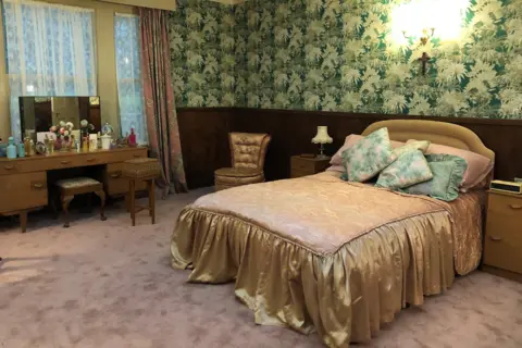 A bedroom with a double bed with gold sheets and green cushions on it, with a dressing table by a window on one side and patterned green wallpaper around the walls