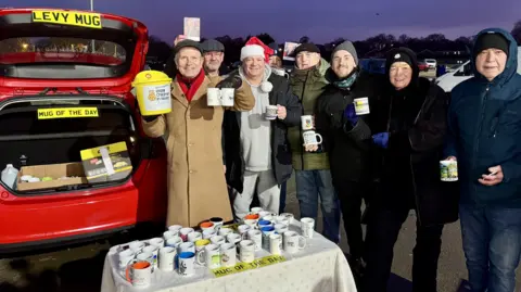 Look North presenter Peter Levy pictured during his mug giveaway in Hull. He is wearing a long brown coat and green cap and is standing next to a red Ford Fiesta with an array of mugs on a table in front of the vehicle. There are several other people in the shot holding mugs.