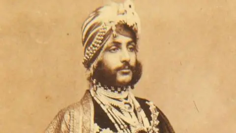 A portrait of Duleep Singh on paper that has aged over time and gone brown. He looks away from the camera and is expressionless. He has 19th Century traditional Indian headwear on as well as clothing. He also has a dark and thick beard. 