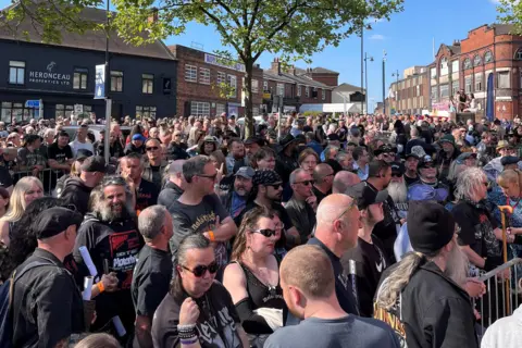A crowd of people gathered in a town square. A tree stands in the middle of the crowd and high street buildings are visible in the background.