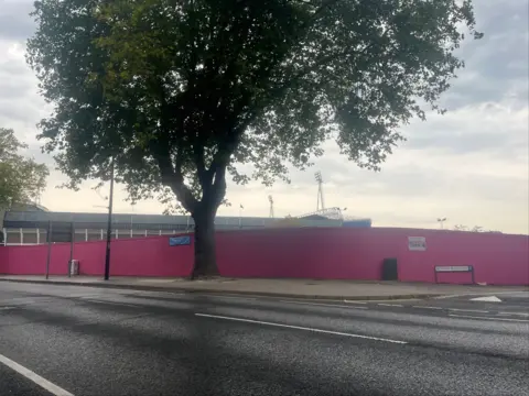 Richard Haugh/BBC Pink wooden hoardings surround a car park. A top of a grandstand and two floodlights are on the horizon beyond the hoardings.