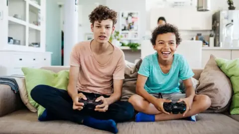Getty Images Two boys with medium-length dark brown hair with games controllers in their hands. One is wearing a pink top and the other wears a light blue top. They are sitting with legs crossed on brown and green cushions. The camera is in the position of the screen. The boy on the right is smiling, the other looks frustrated. They are in a domestic lounge with white furniture.