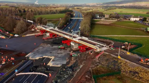 An aerial view of the new bridge being positioned over the M6. It is a light grey with bright red supports underneath rolling it into place.