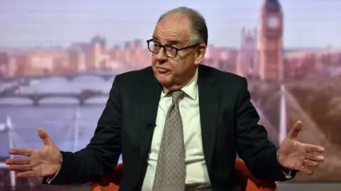 Charlie Falconer pictured in a BBC studio. He is wearing a black suit and a colourful tie. He has black rimmed glasses and short grey hair.