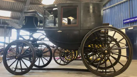 Katie Radley/ BBC A dark coloured historic looking coach with large wheels and a seat on the front of the carriage stands on the floor of a large room with a blue wall to the right and a sign reading exhibition hall.