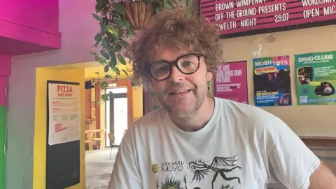 A man with short light brown curly hair and wearing a white T-shirt with a black and yellow design is smiling while sitting in the Future Yard music venue.