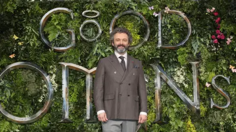 Getty Images  Michael Sheen attends the Global premiere of Amazon Original "Good Omens" at Odeon Luxe Leicester Square on May 28, 2019 in London. He is standing in front of a floral/leafy display with huge silver letters spelling Good Omens attached. Sheen is wearing a grey suit, white shirt and grey patterned tie. He has grey and silver hair and beard. 