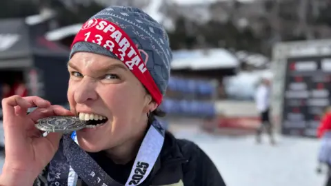 Fran Bullock Fran Bullock biting her medal. She is wearing a hat and is looking at the camera. She has a red banner around the hat which has the word 'Spartan' on it with a number below.