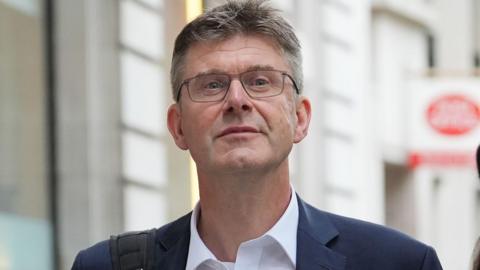Greg Clark wearing a suit and glasses, walking down a street in London