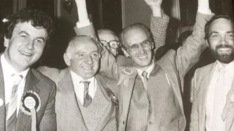 Rachel Stone A black and white picture of Fred Stone is third from the left with Adrian Sanders (L), Wally Dolman, and Michael Mitchell (R) wearing suits and rosettes