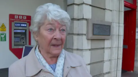 Ruth Mead wearing a light pink coat with a white printed scarf. She is looking to the right. There is a cash machine in the background with a red arrow around it. She is stood in front of a white building with a red door to the right