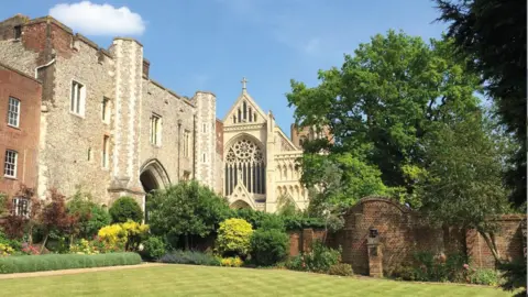 St Albans School showing an ancient brick building, with a church to its side, with trees and a garden. The garden has a wall, with a statue in it, and a large grass lawn.