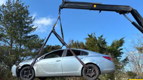 A silver-grey car is being lifted into the air by a small crane. Behind are small trees.