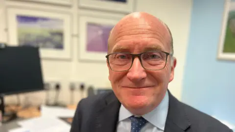 A head-and-shoulders picture of Nick Hulme. He is bald and is wearing glasses, a dark suit, a white shirt and a blue tie with white spots. The background is out of focus, but it appears to be an office.