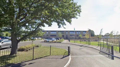 A school is seen from the gates. There is a big green tree next to the gate and the sky is blue.