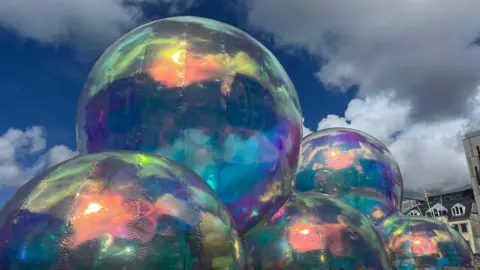 Giant iridescent bubbles that are tall. The sky is blue with white clouds. 