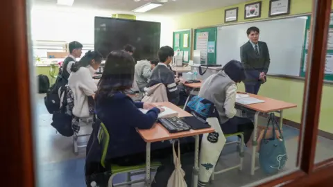 BBC/Hosu Lee A classroom full of students at their desks, using braille materials and magnifiers, while a teacher stands in front of the whiteboard

