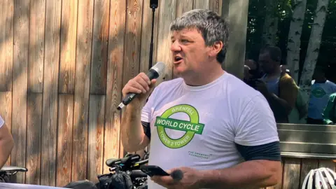 Stephen Aslin wearing a white t-shirt which says Grenfell World Cycle, Tower 2 Tower on it. He is speaking into a microphone and behind him is a wooden fence and his bike leaning against it.