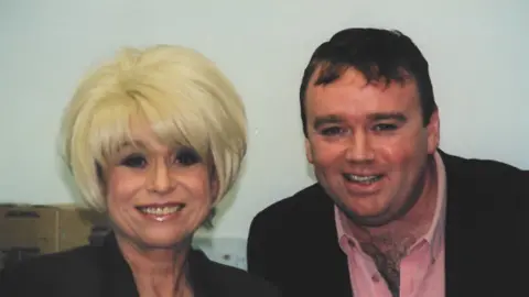 A blonde haired woman in a black top stands next to a man in a black blazer and pink shirt smiling at the camera. 