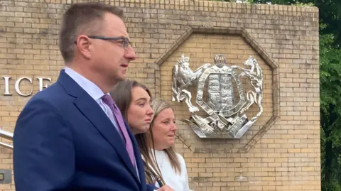 A man with short hair and glasses, wearing a blue suit is in the foreground, standing next to a young woman with long dark hair and a woman dressed in white with long blonde hair. Both women are upset. There are microphones in front of the trio and a court motif on a wall behind them