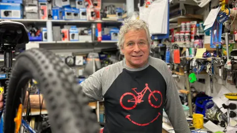 Aaron Lappage is standing in his bike shop. He's a man with curly grey hair. He is wearing a black and grey top with a red bike on it. He is smiling into the camera and leaning on a bike that is elevated, with its back wheel coming into the front of the photo.