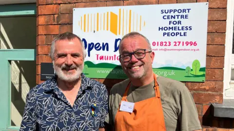 Two men stand side-by-side smiling at the camera in the sun. Simon on the left is wearing a blue open collar shirt, with a white leaf pattern, while Ian wears an orange apron over a khaki-green T-shirt. They are standing in front of the sign for the Open Door building.