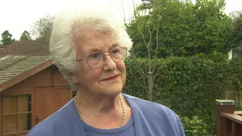 Ann Stribley, who has short white hair, is standing in front of a wooden shed and hedge, and is wearing a gold necklace and is wearing a blue top.