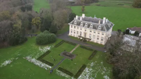A drone view of a large stately home, a cream-coloured rectangular building with a grey roof and four rows of windows, set in formal gardens surrounded by green trees.