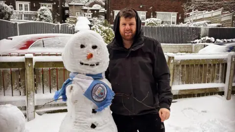 TomMcKeown Tom McKeown stands next to his snowman, which is wearing a light blue Manchester City scarf, in his snow-covered garden. 