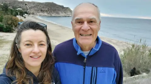 Thompson family A man and woman pose for a selfie, with the background of a sandy bay and a grey-blue sea. The woman is standing to the left, has long brown hair, sunglasses on top of her head and has blue eyes. She is wearing a dark blue hooded top. The man to her right is taller, balding, with grey hair at the sides of his head. He has brown eyes and is wearing a zipped-up fleece in two shades of blue. Both are smiling at the camera. 