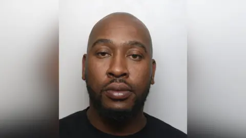 Dedicated Card and Payment Crime Unit Blessing Mudzinge custody image. He is bald and has a dark beard. He wears a black T-shirt.