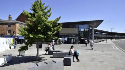An exterior view of Lincoln Central Bus Station. It is a modern-looking glass building. In front of it are concrete benched with people sitting on them and a large tree with green leaves. The sky is blue. 