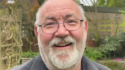Tom Bootyman A close-up photograph taken in a garden of a man with short, thinning hair on the top of the head and a full white beard and moustache. He is wearing rectangular eyeglasses with thin, dark frames. A dark, short-sleeved collared shirt is visible, worn slightly open at the neckline.