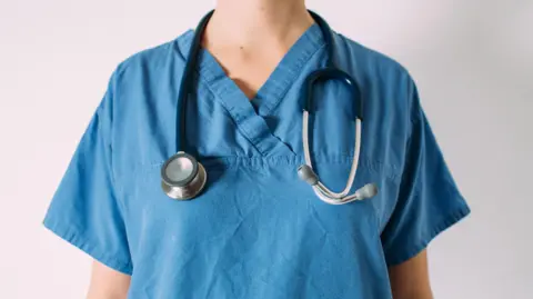 A person is standing facing the camera wearing blue hospital scrubs with a stethoscope around their neck. You cannot see their face or any defining features. 