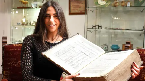 The photograph shows a dark haired woman wearing a black, long-sleeved, mesh-patterned top, holding a large, old book open across her lap. The book has thick, yellowed pages. The open pages display printed text arranged in columns. There is also a framed certificate hanging on the wall and glass display cabinets.