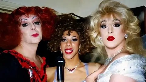 Vixon Herts Three drag artists singing on stage. On the left is Regina, with a dark red wig on, wearing heavy black eyeliner, mascara and bright red lipstick, a frilled v-neckline top - the v neck is decorated with red ruffles. In the middle is Lucia, with red lipstick, a short curly bobbed hair and a blue strappy top, and Vixen has blonde bouffant hair, a white dress, diamond earrings and necklace and bright red lipstick. This is a head and shoulder shot. 