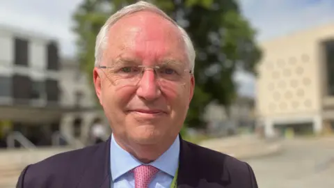 Chris is wearing a suite and tie. He is standing outside on a sunny day smiling into the camera. He has short white hair and light coloured eyes.