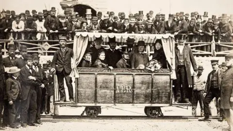 The Regency Society A black and white image of a large group of men. There is a handful of men inside a carriage on the Volk's Railway. 