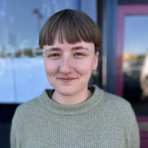 A woman with dark hair and a greyish/green jumper stands in front of a cafe