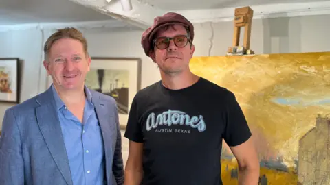 Grant Ford smiles at the camera, wearing blue shirt and jacket. Next to him is Asa, who wears glasses, a peaked brown hat and a black t-shirt. They stand in front of White's last work.