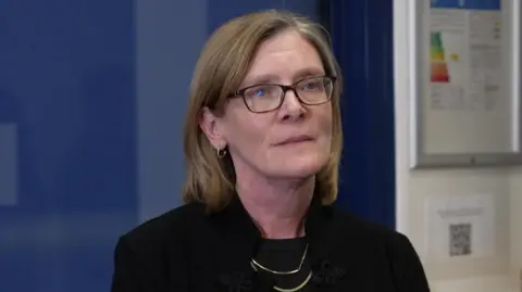 A woman wearing a black blazer and black top. She is wearing glassed and a gold necklace and has shoulder length hair. There is a blue and white wall behind him. 