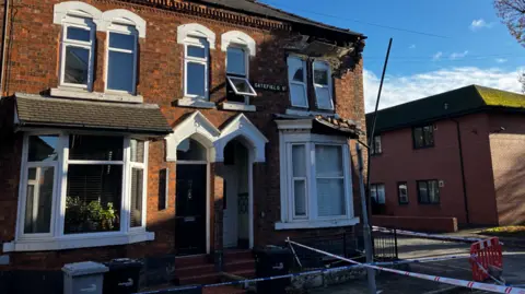 BBC An end terrace has bricks on top of the window frame, and there is damage above the upper windows. There is a fire service tape sealing off the road around the property