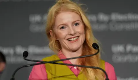 Hannah Spencer has strawberry blonde hair, a pink blouse and and green waistcoat