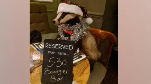 The stuffed badger Bertie wearing a Christmas hat and some tinsel around its head. It is at a pub table with a sign saying reserved for the Badger Bar staff party.