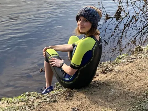Sheila Mitchell A young woman is sitting on the edge of some water. She is wearing a green and black wetsuit and a woolly hat. She is looking back over her shoulder and smiling at the camera.