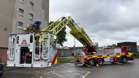 View of the new turntable ladder a firefighter is in the basket in the foreground which is being lifted off the ground by the fire engine 