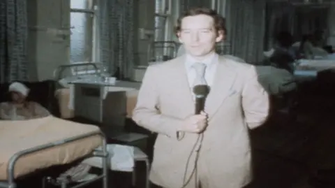 Michael Buerk reporting from a hospital ward the day after the bombings. He is standing facing the camera with a microphone in his hand, and is wearing a beige suit with a striped tie. There are beds behind him, and to the right is a patient with most of their head and face obscured by bandages.