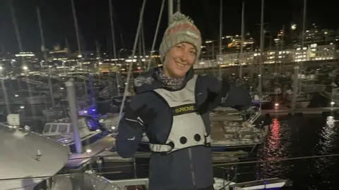 Jeremy Mattison Nina Mattison stood with her running gear on by the dock. She is smiling at the camera and is wearing a woolly hat. 