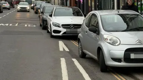 A row of around seven cars parked badly on double yellow lines, overlapping onto the pavement at Southgate Street, Gloucester. Their number plates have been blurred out.