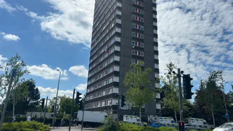 BBC A multi-storey block of flats, which features brown stone striped with red around windows. A number of emergency services vehicles are parked at the base of the tower. The sky is blue, with high clouds.